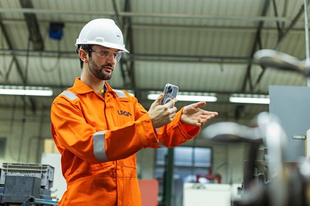 Ingeniero con equipo de seguridad utilizando un dispositivo móvil para inspeccionar equipos industriales en una planta de fabricación.